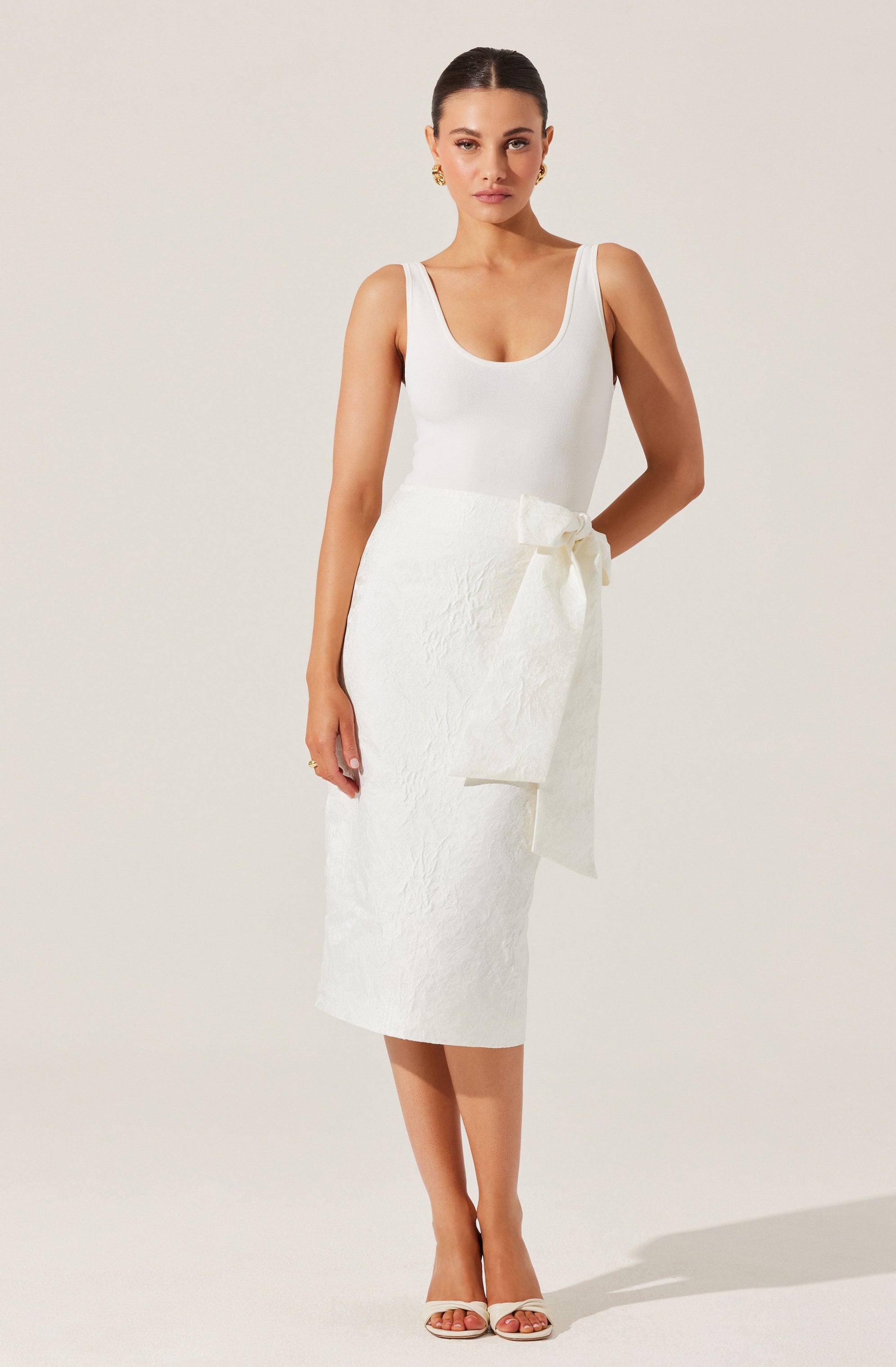 A woman is wearing a sleeveless white top with wide shoulder straps and a scoop neckline, paired with a high-waisted, mid-length white skirt featuring a textured fabric and a large bow tied at the waist. She is also wearing white open-toe high heels and gold earrings.
