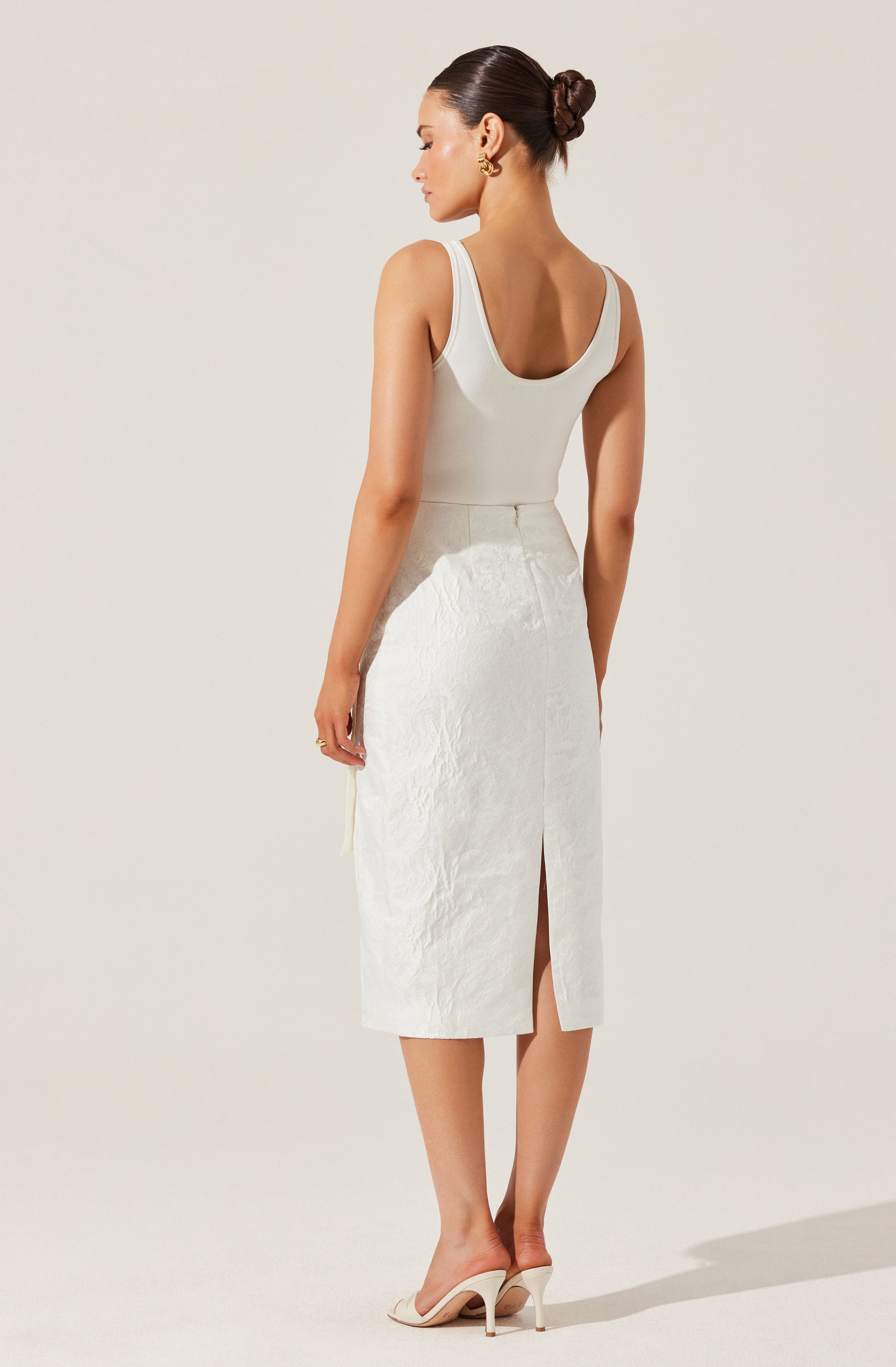 A woman is wearing a sleeveless, fitted, white top with a scoop neckline, paired with a high-waisted, mid-length white skirt that has a front slit. She is also wearing light-colored high-heeled shoes and gold hoop earrings. Her hair is styled in a polished, low bun.