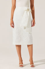 A woman is wearing a white, textured, midi-length skirt with a large side bow tied at the waist. She is also wearing white open-toe high heels.