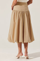 A long beige skirt with a high waist and gathered fabric that creates a voluminous, bubble-like effect at the hem. The skirt is paired with a light-colored top and white high-heeled shoes.