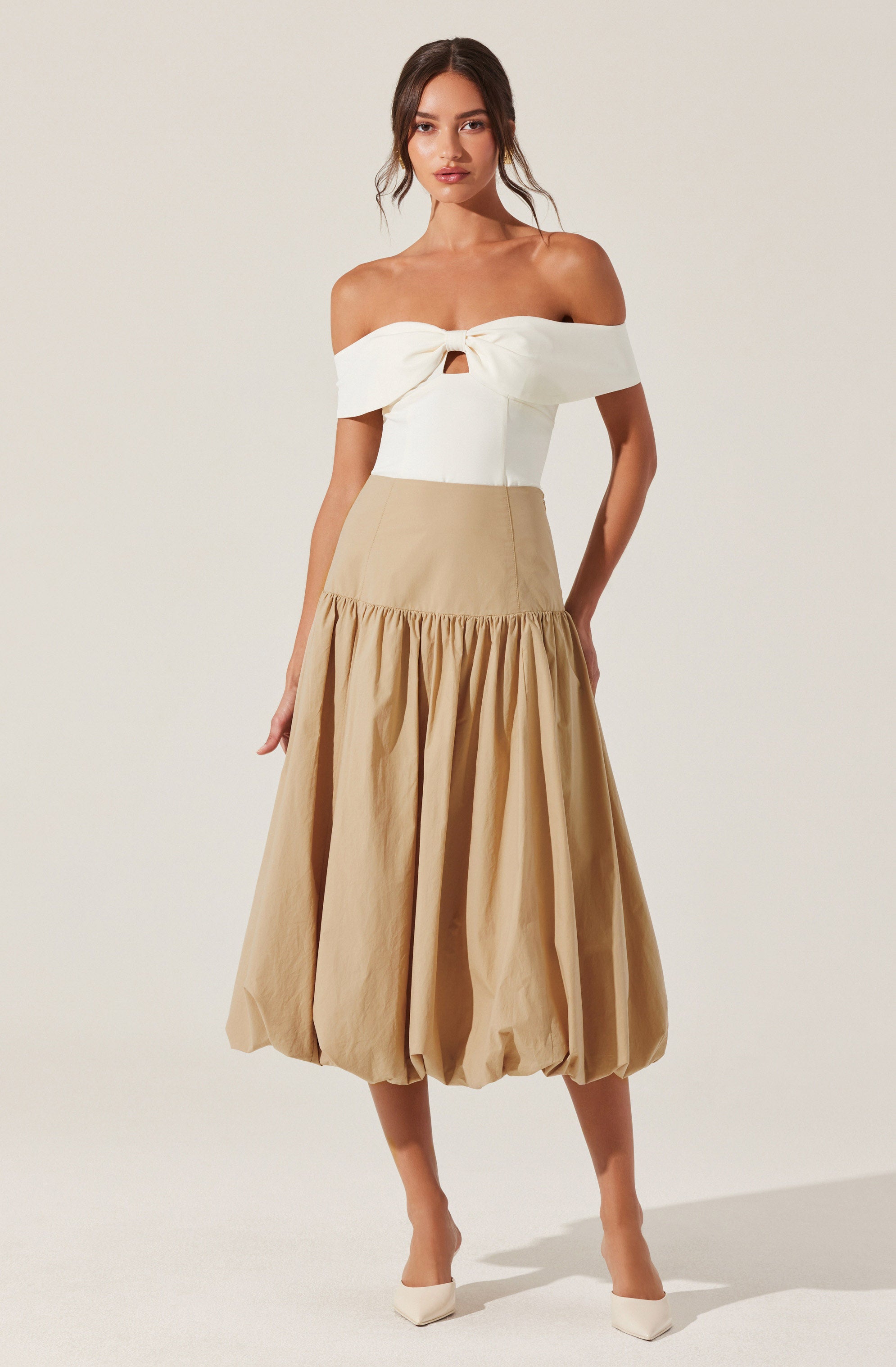 A woman is wearing an off-the-shoulder white top with a front knot and a small cutout below the knot. She pairs it with a high-waisted, beige, balloon-style skirt that has a gathered, voluminous design at the hem. She completes the outfit with white pointed-toe shoes.