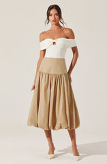 A woman is wearing an off-the-shoulder white top with a front knot and a small cutout below the knot. She pairs it with a high-waisted, beige, balloon-style skirt that has a gathered, voluminous design at the hem. She completes the outfit with white pointed-toe shoes.