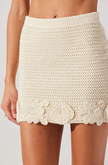 A woman is wearing a cream-colored crocheted skirt with a wide ribbed waistband. The skirt features a textured, open-knit pattern throughout and is decorated at the hem with large, intricate crocheted floral motifs.