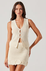 A woman is wearing a sleeveless, cream-colored, knitted outfit consisting of a fitted top with a deep V neckline and a matching skirt. The top features a front opening with a scalloped edge, and the skirt has a decorative floral crochet pattern near the hem. The knit pattern appears to be slightly open, creating a textured look.