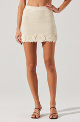 A woman is wearing a cream-colored, crochet mini skirt with a textured pattern. The skirt features decorative crochet flower appliqués at the hemline, adding a floral detail to the garment. She pairs the skirt with strappy, open-toe sandals. Her top is not visible in the image.