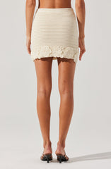 A person is wearing a cream-colored, textured knit skirt with a floral crochet detail at the hem. The skirt is high-waisted and fitted, ending above the knees. They are also wearing open-toe, black heeled sandals.