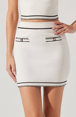 A woman is wearing a white, fitted skirt and matching crop top, both featuring black trim accents. The skirt has two decorative pockets with black trim and gold button details. The crop top has a black trim along the hem and a subtle black line detail around the waist.