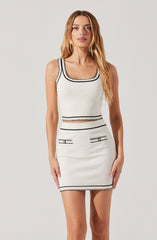 A woman is wearing a sleeveless, fitted white dress with black borders and decorative stitching. The dress features a square neckline and a sheer horizontal cutout at the waist. It has two small front pockets with gold-colored button details and is accessorized with matching black trim along the hemline, neckline, armholes, and around the waist cutout.