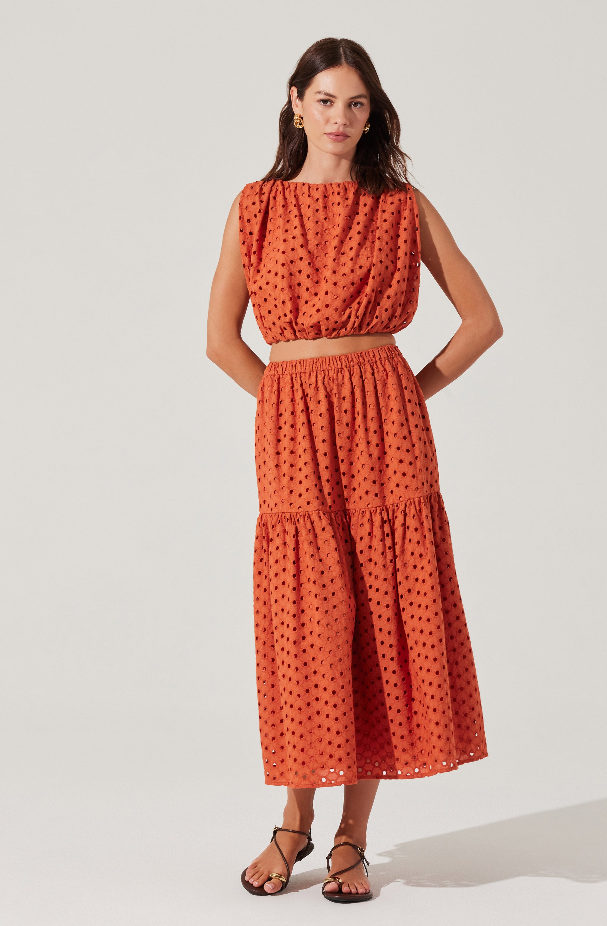 A woman is wearing a sleeveless, orange two-piece outfit made of a fabric with a black polka dot pattern. The top is a loose, cropped blouse with a gathered neckline and armholes. The skirt is high-waisted, flowy, and midi-length, featuring a tiered design with gathered fabric at the seams.