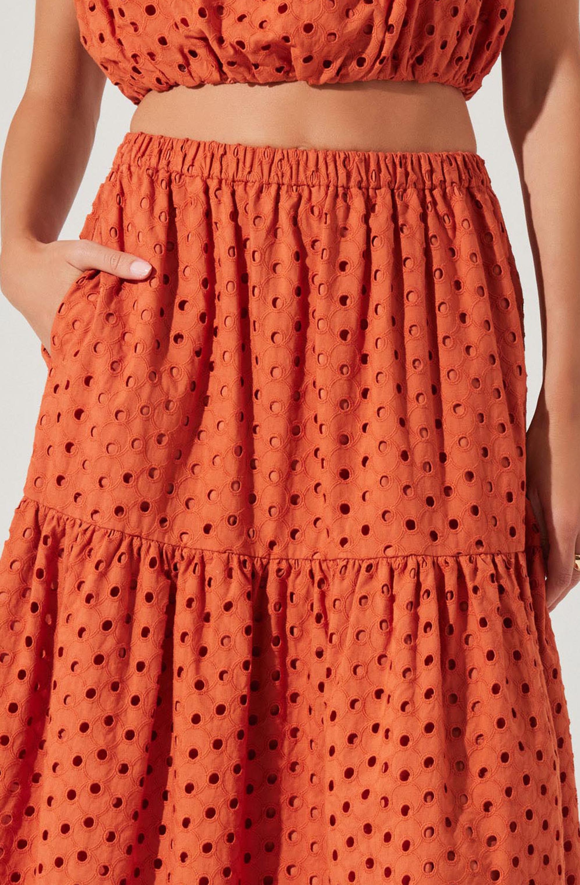 A woman is wearing a matching two-piece set made of orange fabric with a pattern of evenly spaced, circular cutouts. The top is a sleeveless crop with an elasticized band at the hem, and the skirt is a high-waisted, tiered design with an elastic waistband and gathered fabric at the tiers. The skirt has large side pockets, and the woman's hand is resting in the pocket on her right side.
