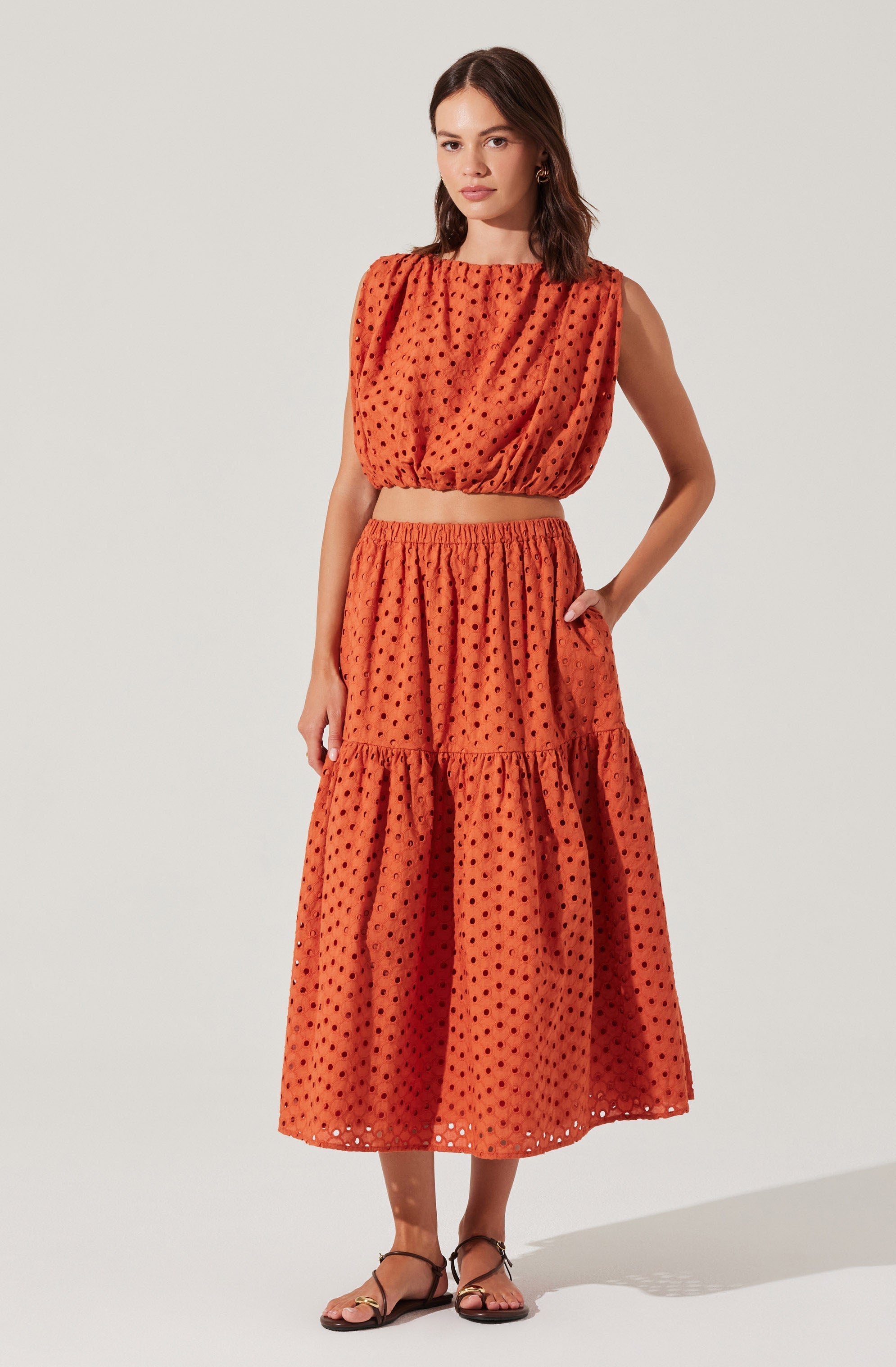 A woman is wearing a two-piece outfit made of orange fabric with a black polka dot pattern. The top is a sleeveless, loose-fitting blouse with a round neckline and gathered fabric at the shoulders, creating a slightly puffed effect. The skirt is a long, tiered midi length with an elastic waistband, and it features a ruffled tier near the hem. She is also wearing black strappy sandals.