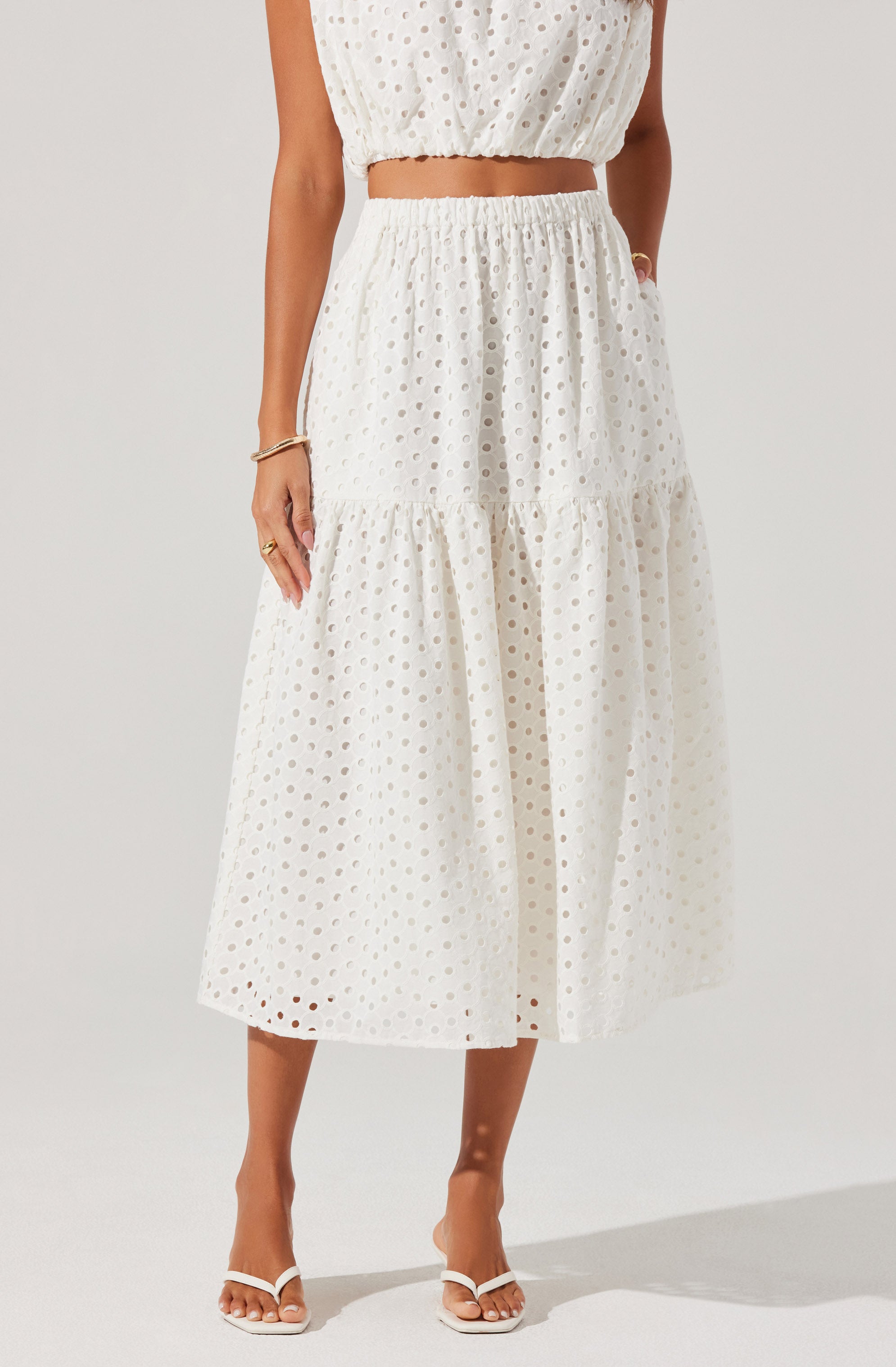 A woman is wearing a white two-piece outfit with eyelet embroidery. The top is a sleeveless, cropped style with an elastic band at the hem, and the midriff is exposed. The skirt is high-waisted, with an elastic waistband, and has a tiered design with gathered fabric creating a flowing silhouette that falls to mid-calf length. She is also wearing white strappy high-heeled sandals.