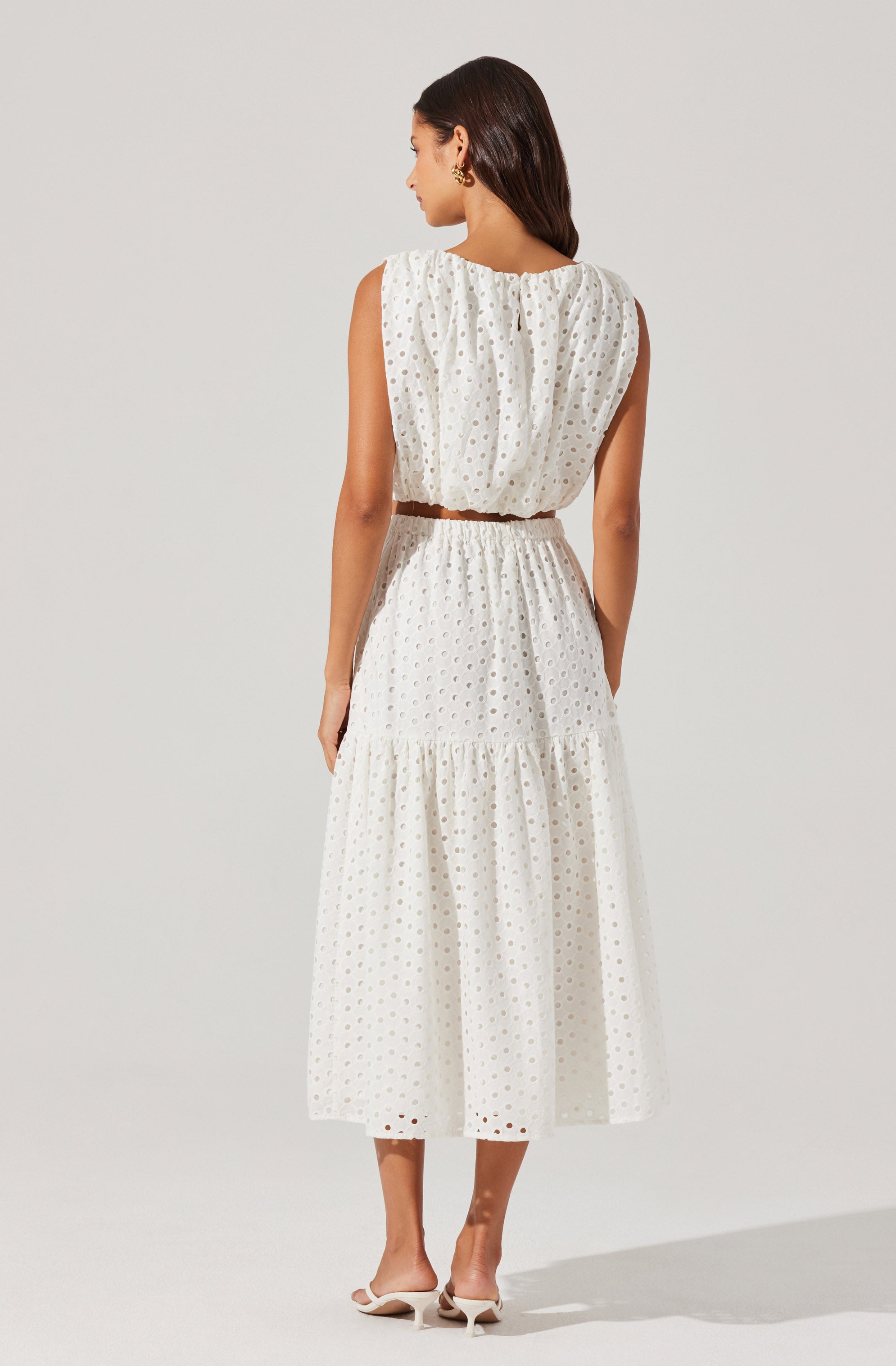 A woman is wearing a sleeveless, white dress with an eyelet pattern. The dress features a fitted waistline with a drawstring or elastic gathering, a gathered neckline, and a full, ankle-length skirt with a tiered design. She accessorizes with gold earrings and white pointed-toe heels with a small heel.