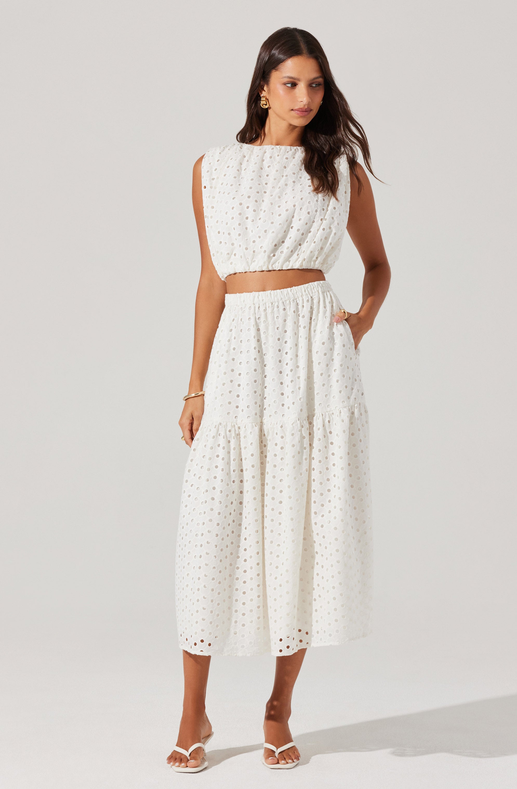 A woman is wearing a matching sleeveless white crop top and a high-waisted, ankle-length skirt. The fabric of both pieces features a subtle eyelet pattern with small circular cutouts. The top is loose-fitting and gathered at the waistband, while the skirt has a tiered design with a gathered seam around the midsection. She accessorizes with gold hoop earrings, a bracelet, and white open-toe sandals.