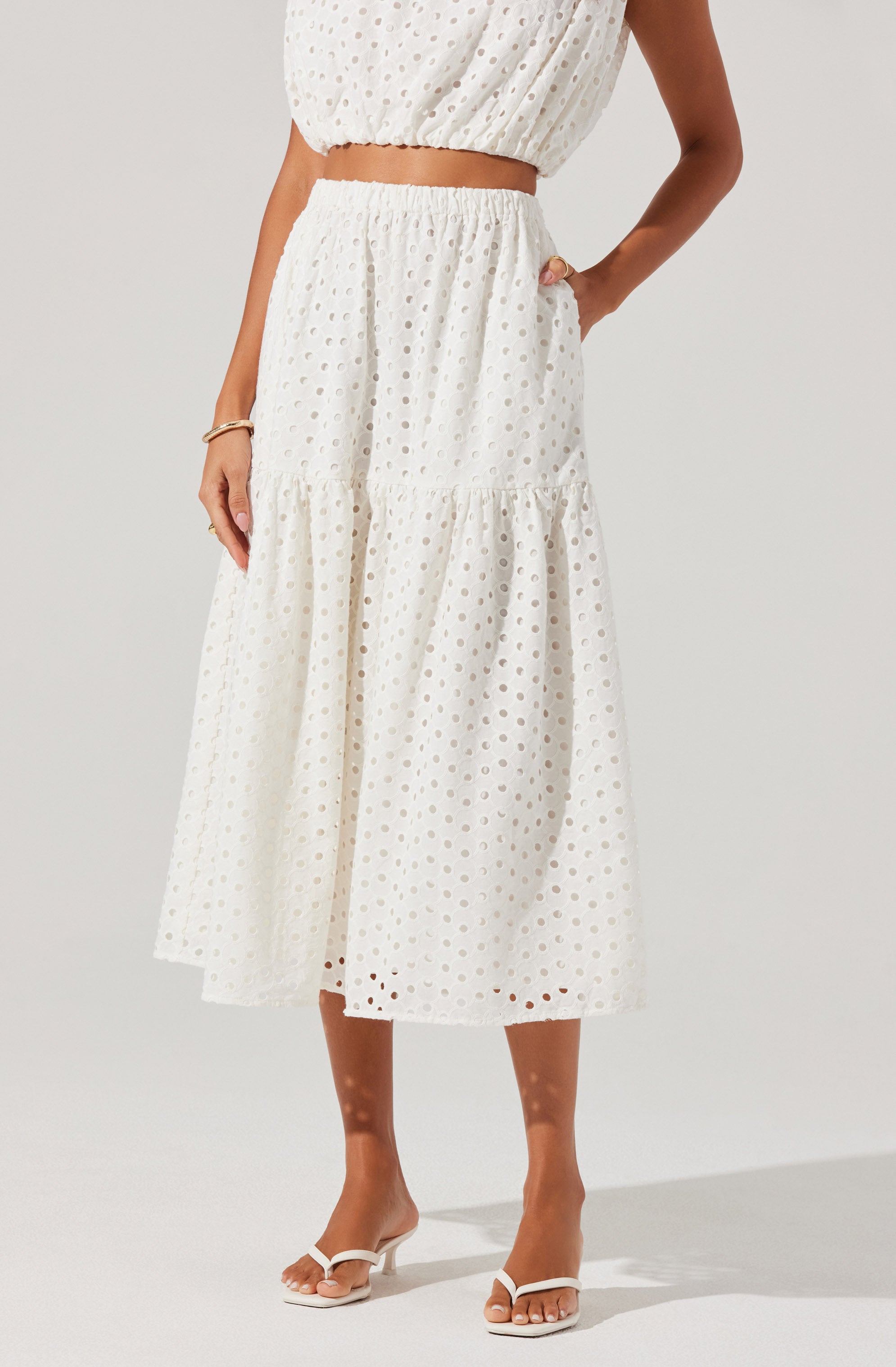 A woman is wearing a matching white two-piece outfit made of eyelet fabric. The top is a sleeveless cropped blouse with a loose fit, and the skirt is a high-waisted, ankle-length skirt with a tiered design, featuring a gathered seam near the hem. She is also wearing white heeled sandals and accessorized with a bracelet on her left wrist.