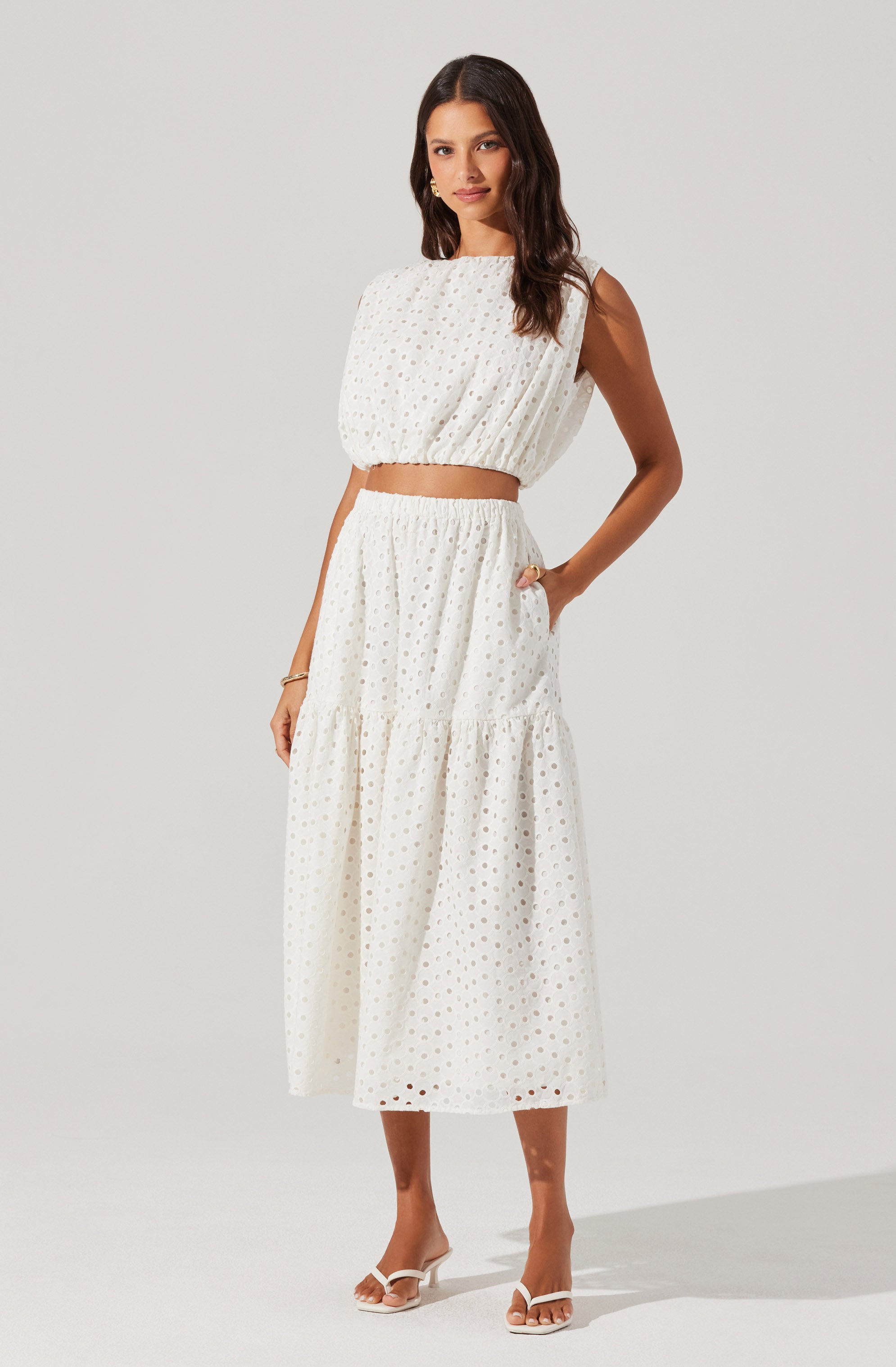 A woman is wearing a sleeveless, two-piece white outfit with a cropped top and a high-waisted, tiered maxi skirt. The fabric has a subtle eyelet pattern with small cutouts throughout. The cropped top is loose-fitting with a rounded neckline, and the maxi skirt is flowy with a gathered seam at the tier.