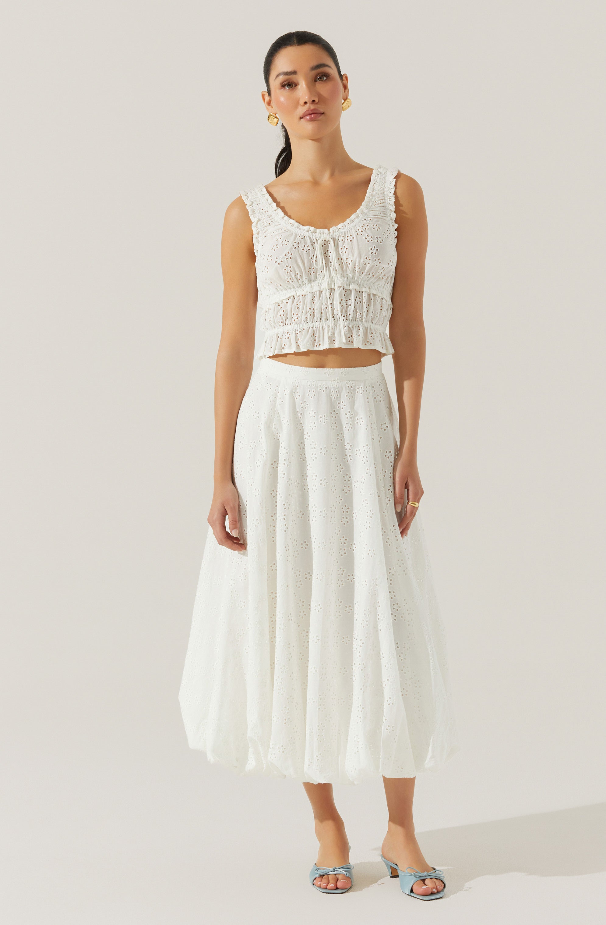 A woman is wearing a sleeveless, white, eyelet lace crop top with ruffled details and a rounded neckline. She pairs it with a high-waisted, white, ankle-length skirt made of eyelet fabric with a subtle pattern of small holes. She completes her look with light blue, open-toe, heeled sandals featuring a bow detail on the front. She is also wearing gold hoop earrings and a gold ring on her right hand.