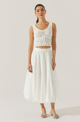 A woman is wearing a sleeveless, white, eyelet lace crop top with ruffled details and a rounded neckline. She pairs it with a high-waisted, white, ankle-length skirt made of eyelet fabric with a subtle pattern of small holes. She completes her look with light blue, open-toe, heeled sandals featuring a bow detail on the front. She is also wearing gold hoop earrings and a gold ring on her right hand.