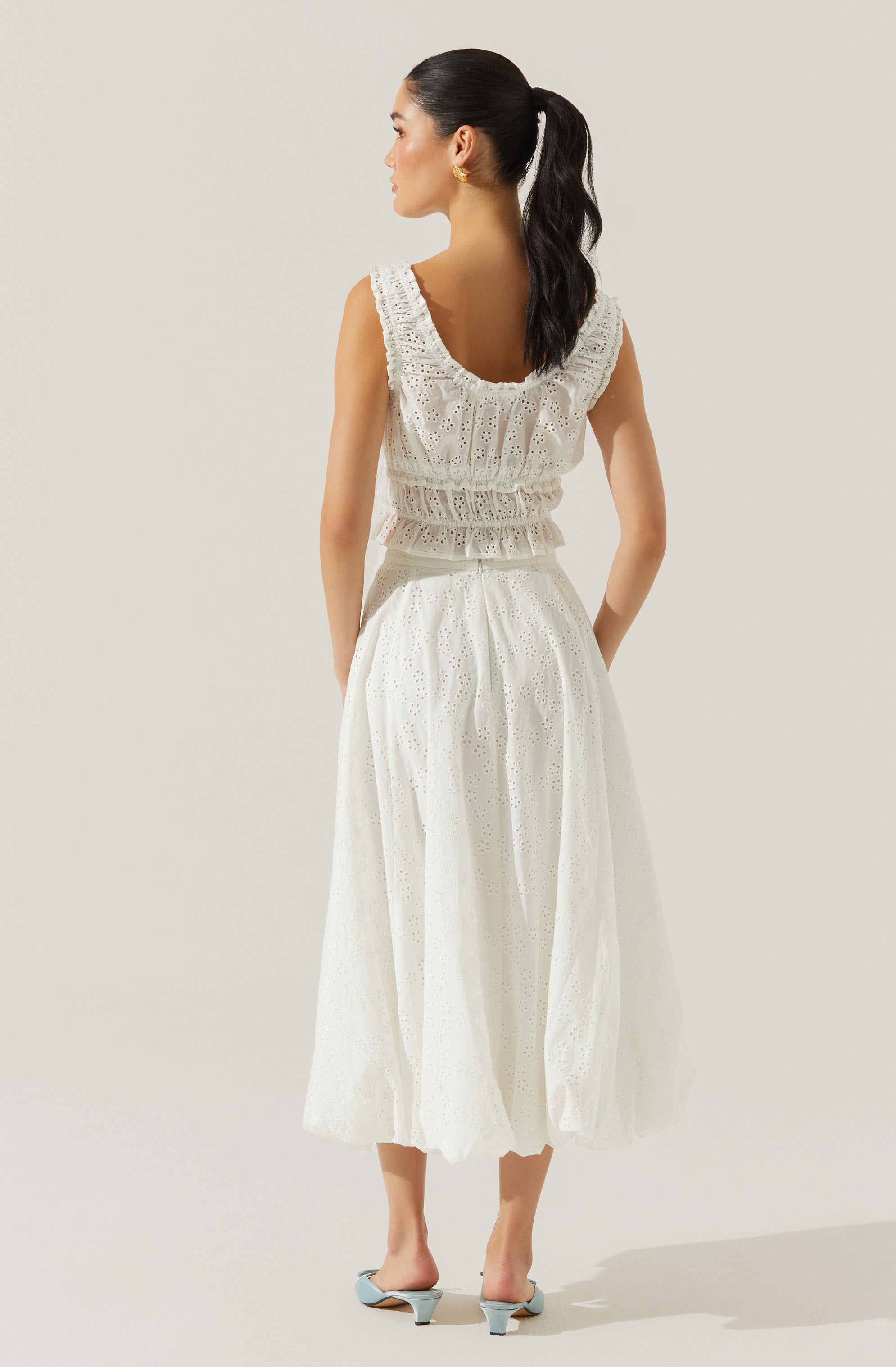 A woman is wearing a sleeveless, white eyelet dress with ruffled straps and a ruffled, fitted waist. The dress has a full, ankle-length skirt with intricate eyelet detailing. She is also wearing light blue, open-toe, low-heeled sandals.
