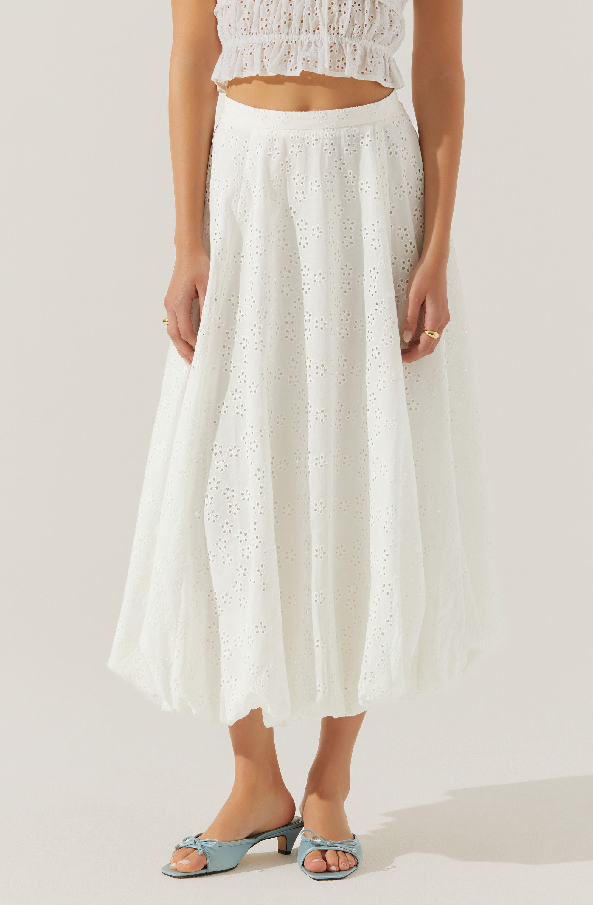 A woman is wearing a white, eyelet, ankle-length skirt with a flowy fit. She pairs it with a matching white eyelet crop top with a ruffled hem. She is also wearing light blue open-toe sandals with a small heel and a bow detail on the front.