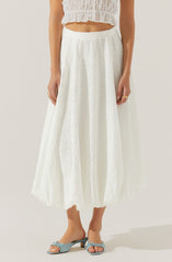 A woman is wearing a white, eyelet, ankle-length skirt with a flowy fit. She pairs it with a matching white eyelet crop top with a ruffled hem. She is also wearing light blue open-toe sandals with a small heel and a bow detail on the front.