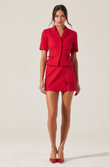 A woman is wearing a matching red short-sleeve blazer and skirt set. The blazer features a notched collar, buttons down the front, and decorative ties at the waist. The skirt has a front slit, and a bow detail at the hem. She is also wearing red high-heeled shoes.