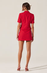A woman is wearing a matching red blazer and mini skirt set. The blazer has short sleeves and a tailored fit. The mini skirt is fitted and ends above the knees. She is also wearing red high-heeled shoes with ankle straps. Her hair is styled in a loose bun with some strands framing her face.