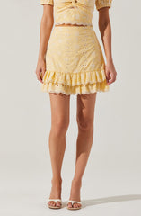 A woman is wearing a light yellow two-piece outfit consisting of a short-sleeved, eyelet lace crop top with scalloped edges and a matching high-waisted skirt with ruffled hem and eyelet lace details. She is also wearing white open-toe sandals.
