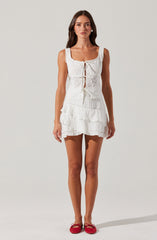 A woman is wearing a sleeveless white dress with a fitted bodice that features a lace trim around the neckline and armholes, and a front keyhole opening with a tie closure. The dress has embroidered and eyelet details, with a tiered, ruffled skirt that reaches mid-thigh. She is also wearing red ballet flats with small bows on the front.