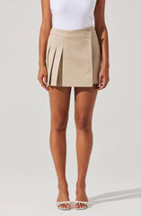 A woman is wearing a beige skirt with pleats on the left side and a plain panel on the right side. The skirt has a fitted waistband. She is also wearing a white sleeveless top, white open-toe high-heeled sandals, and a ring on her left hand.