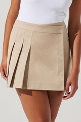A woman is wearing a beige mini skirt with pleats on the left side. The skirt has a straight waistband and appears to be made of a lightweight fabric. She is also wearing a plain white sleeveless top tucked into the skirt.