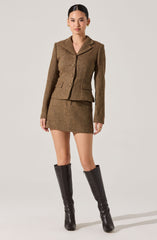 A woman is wearing a brown, textured, tailored blazer with a notched lapel collar, long sleeves, and three buttons down the front. She pairs it with a matching short skirt. She is also wearing tall, black leather boots that reach just below the knee.