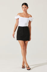 A woman is wearing an off-the-shoulder white top with puffed sleeves and a black high-waisted, above-the-knee skirt. She has on black open-toe heels with thin straps.