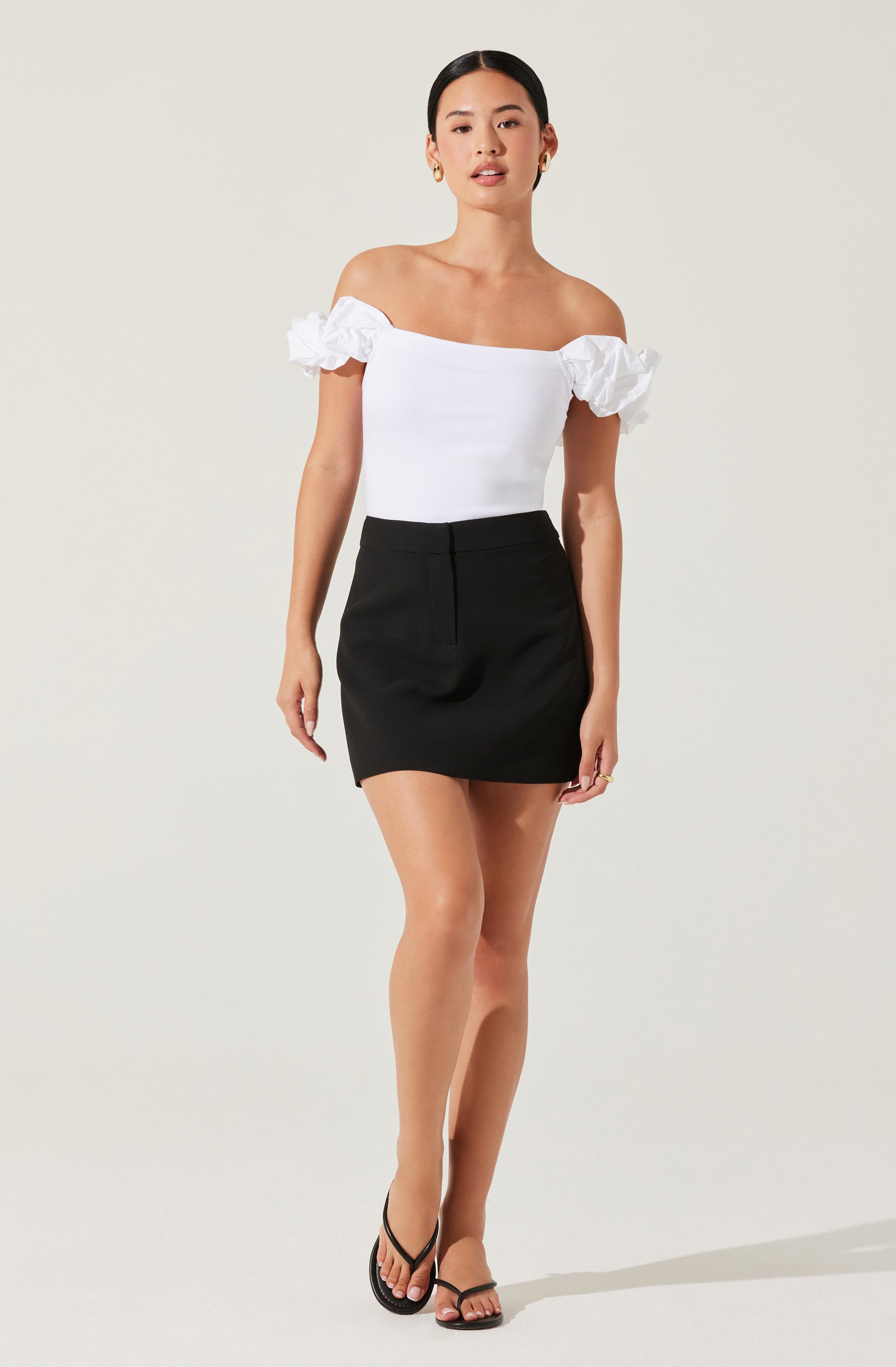 The woman is wearing an off-the-shoulder white top with ruffle details on the sleeves, paired with a high-waisted black mini skirt. She is also wearing black strappy high-heeled sandals.