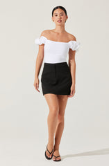 The woman is wearing an off-the-shoulder white top with ruffle details on the sleeves, paired with a high-waisted black mini skirt. She is also wearing black strappy high-heeled sandals.