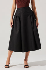 A woman is wearing a high-waisted, wide-leg black skirt with pleats and gathered fabric details on the sides. She pairs it with a sleeveless, fitted, rust-colored top with a wraps design. She is also wearing black open-toed heels with thin straps and a simple gold bracelet on her right wrist.