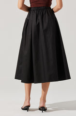 A woman is wearing a high-waisted, ankle-length black skirt with a full, voluminous silhouette. The skirt features an elastic waistband and large front pockets. She is also wearing a fitted, sleeveless top in a dark red or burgundy color.