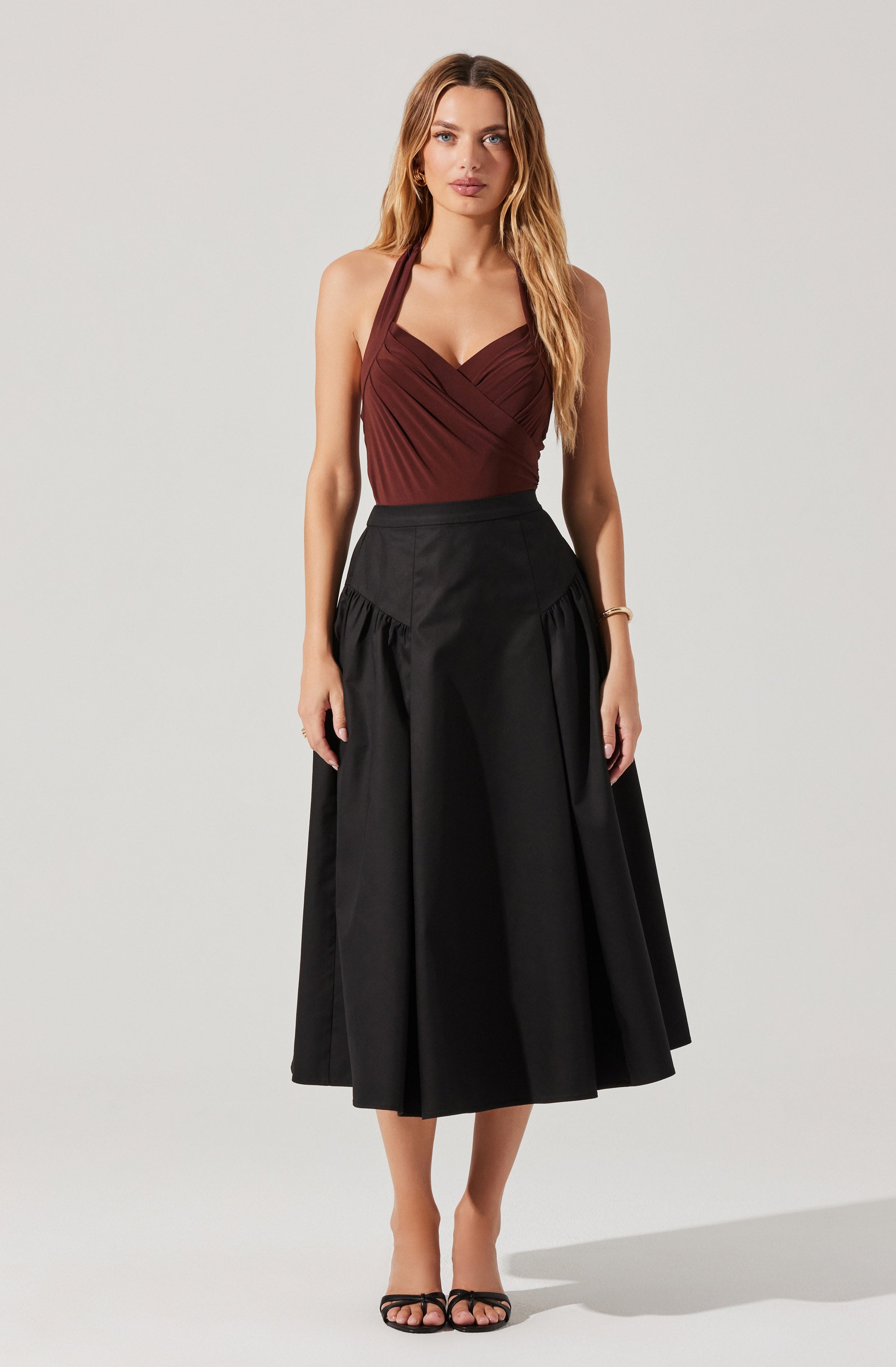 A woman is wearing a sleeveless, burgundy halter top with gathered detailing at the bust, paired with a high-waisted, black midi skirt that features a full, pleated design. She is also wearing black strappy high-heeled sandals.