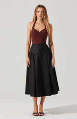 A woman is wearing a sleeveless, burgundy halter top with gathered detailing at the bust, paired with a high-waisted, black midi skirt that features a full, pleated design. She is also wearing black strappy high-heeled sandals.