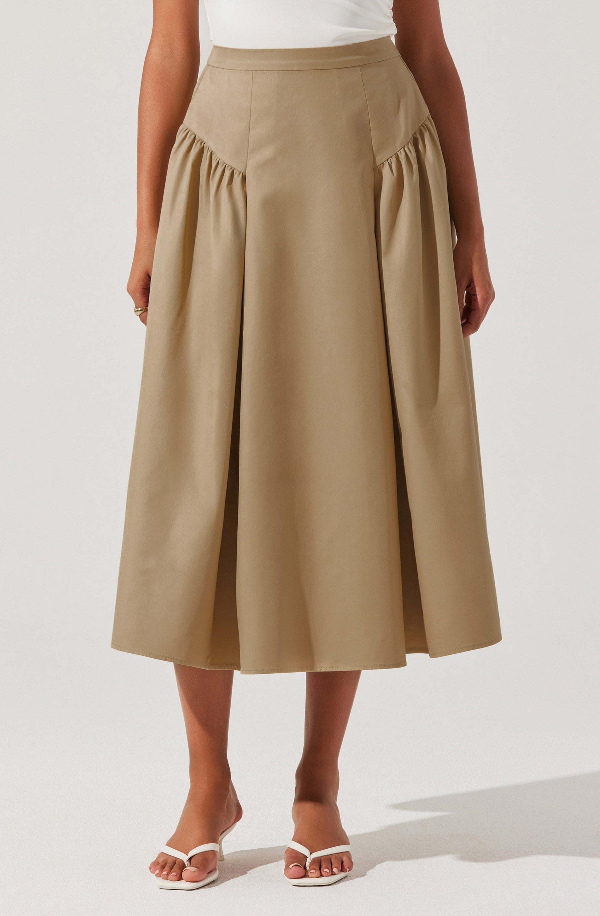 A woman is wearing a high-waisted, beige midi skirt with a full, gathered design on the sides. The skirt is paired with a white top tucked in and white open-toed heels.