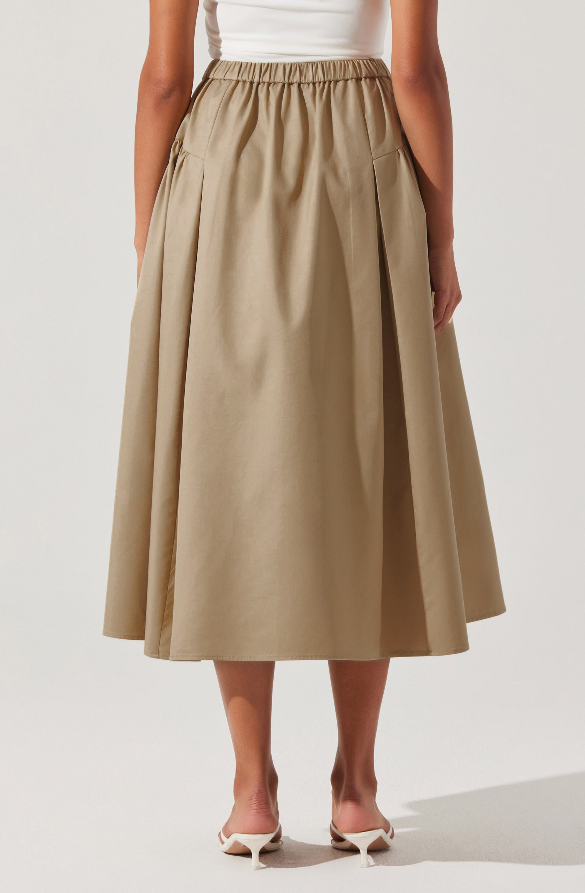 A woman is wearing a beige midi-length skirt with a gathered elastic waistband and large patch pockets on the front. She is also wearing a sleeveless white top and white pointed-toe heels.
