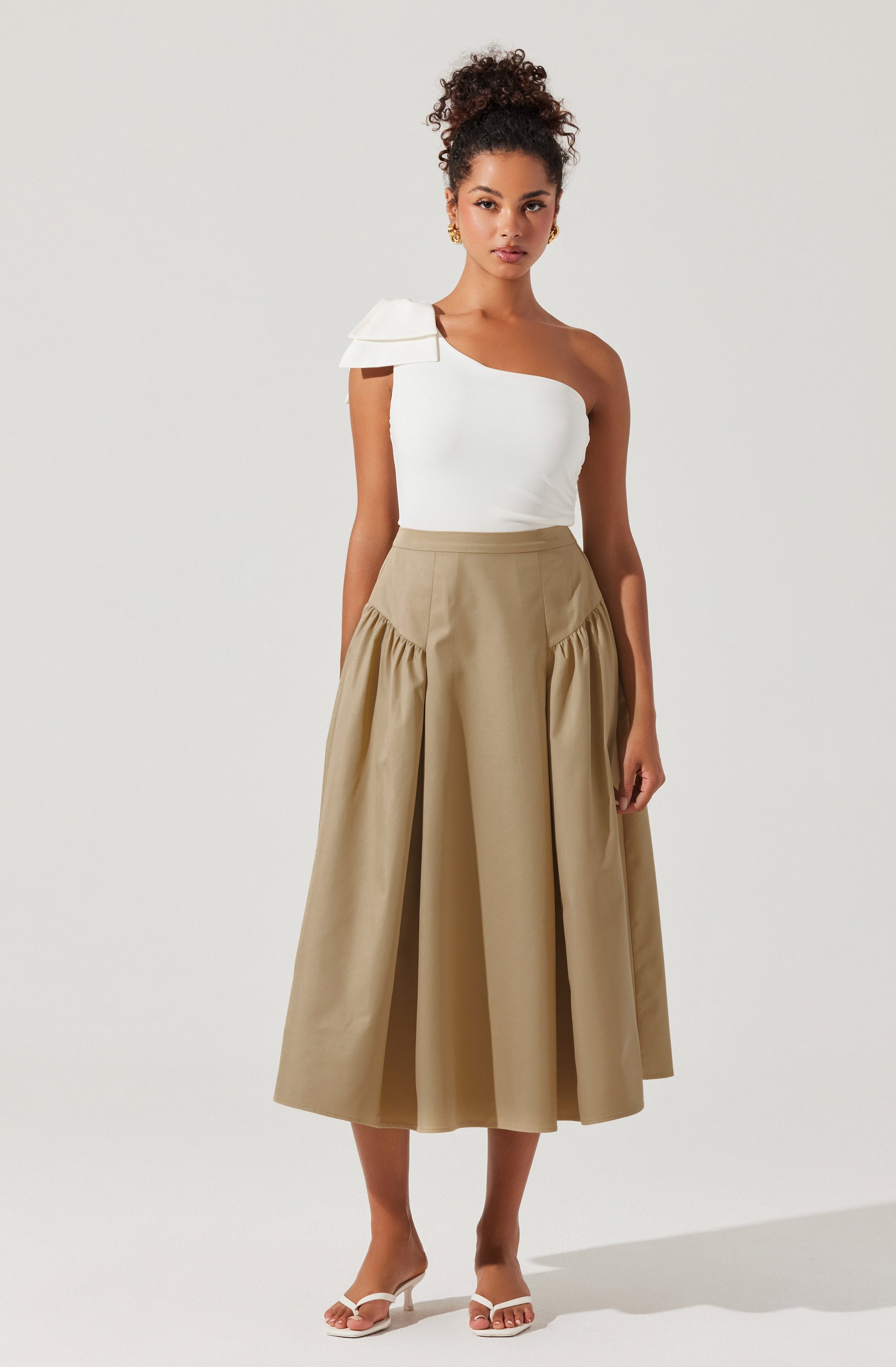 The woman is wearing a one-shoulder white top with a large decorative shoulder detail on the left side, paired with a high-waisted, beige, midi-length skirt featuring gathered fabric panels on the sides. She is also wearing white open-toed high-heeled sandals.