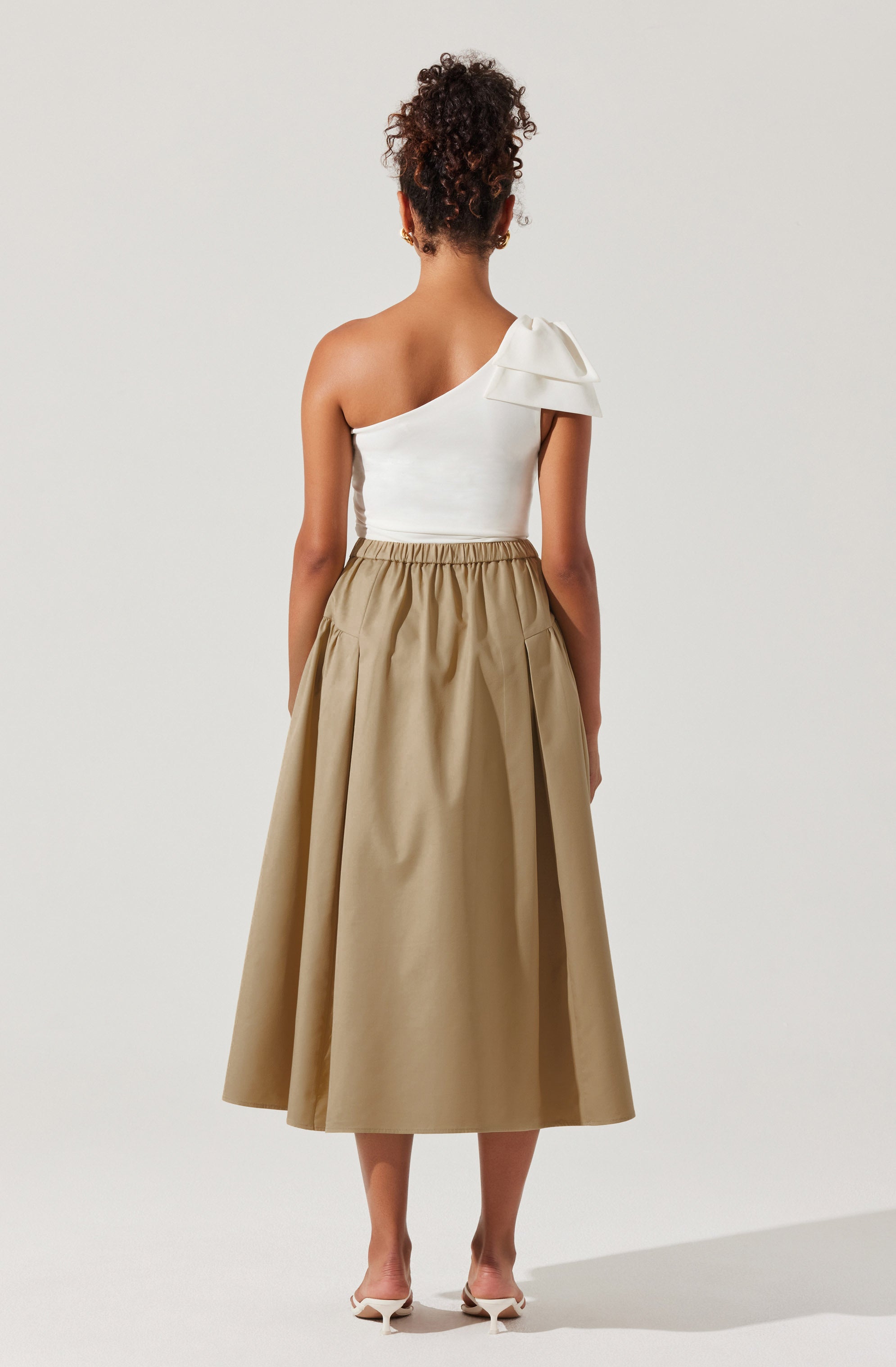 A woman is wearing a one-shoulder white top with a large ruffle detail on the shoulder, paired with a high-waisted, beige, A-line midi skirt with pleats and elastic waistband. She is also wearing beige high-heeled shoes.