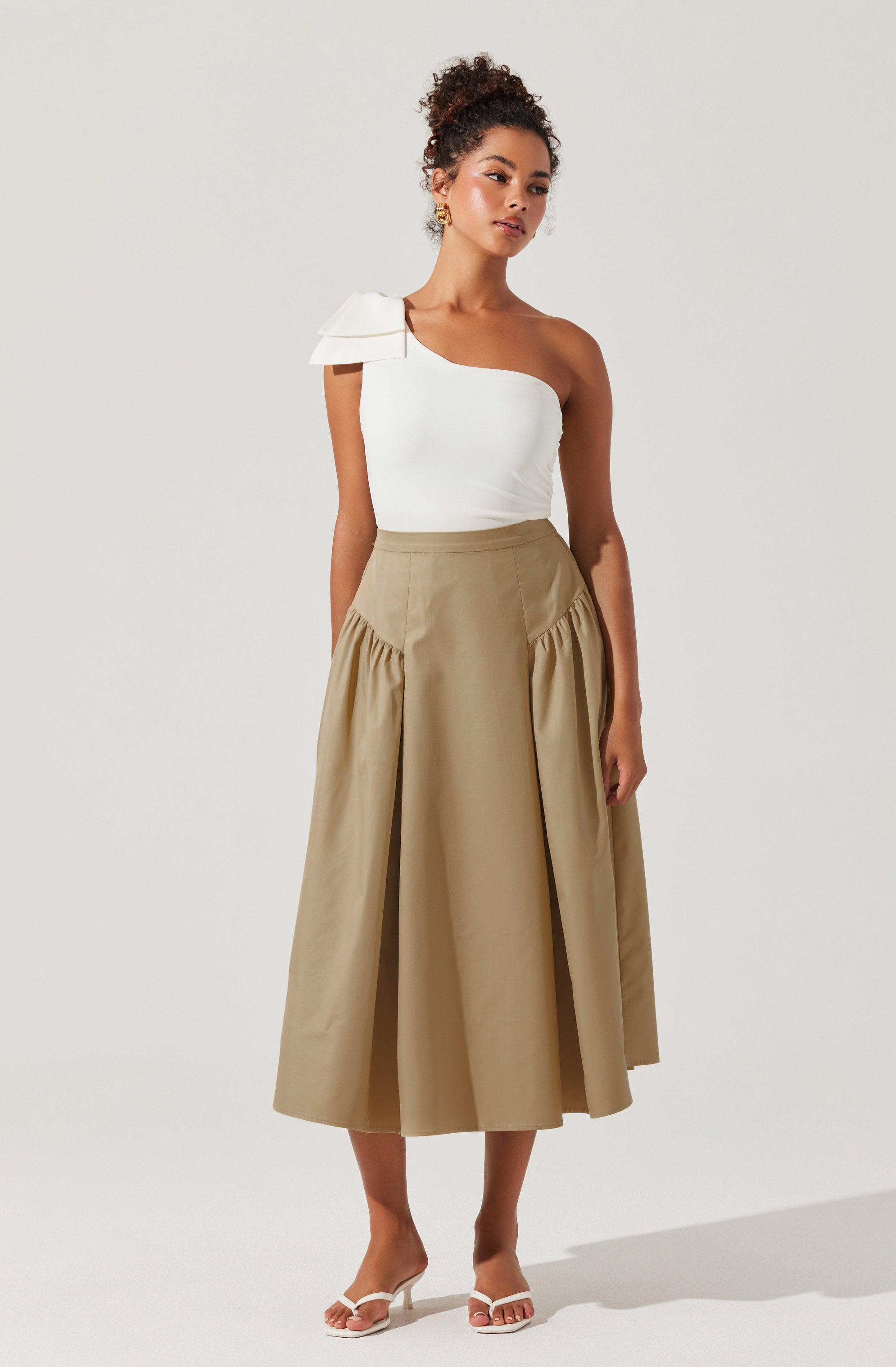A woman is wearing a white, one-shoulder top with a large, decorative shoulder ruffle on the left side. She pairs it with a high-waisted, beige, A-line midi skirt that features gathered fabric details at the hips. She completes the outfit with white strappy heeled sandals and gold earrings.