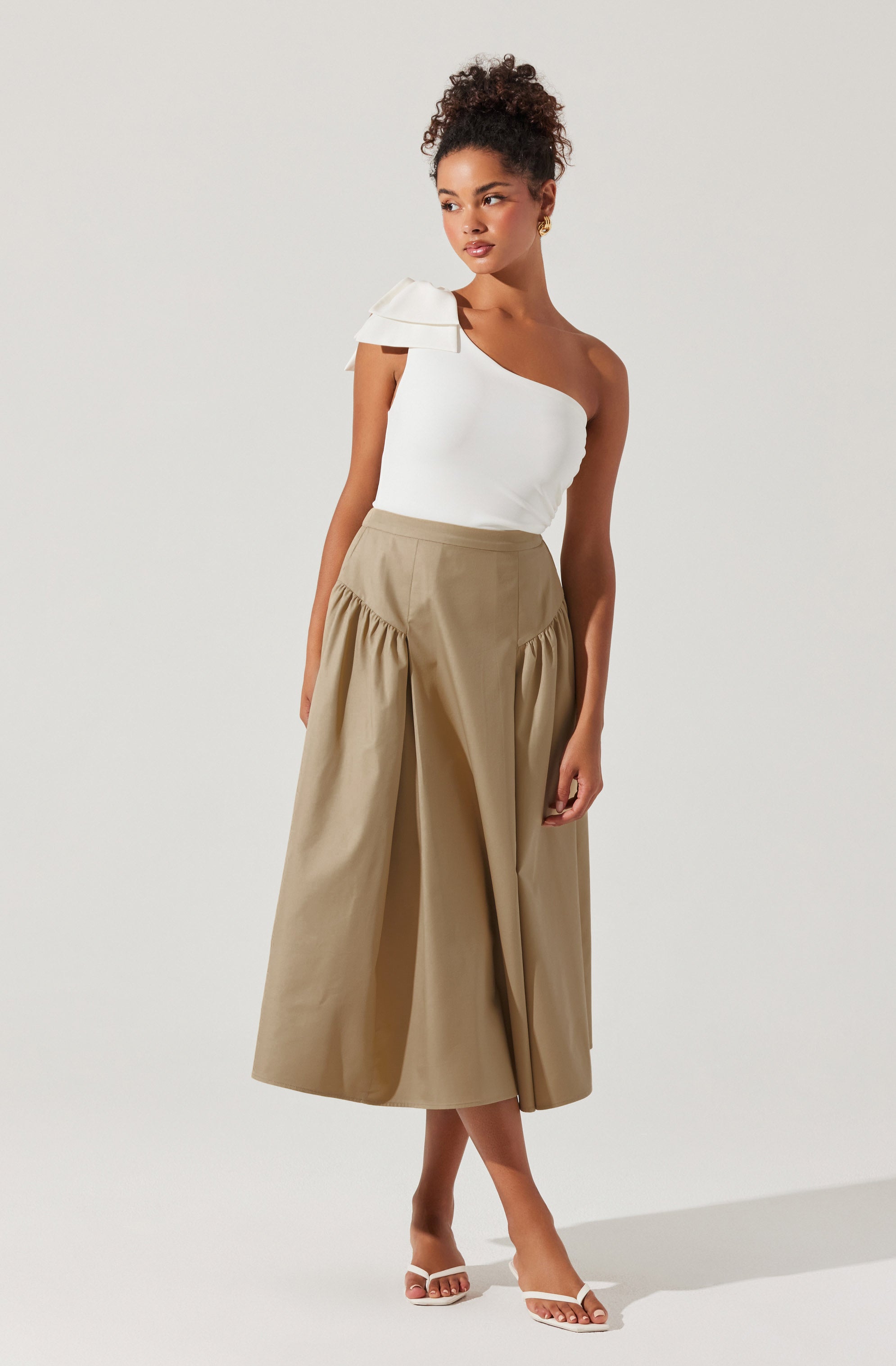 A woman is wearing a white, one-shoulder top with a decorative ruffle on the shoulder, paired with a high-waisted, beige, wide-leg cropped pant with gathered details at the hips. She is also wearing white open-toe heeled sandals.