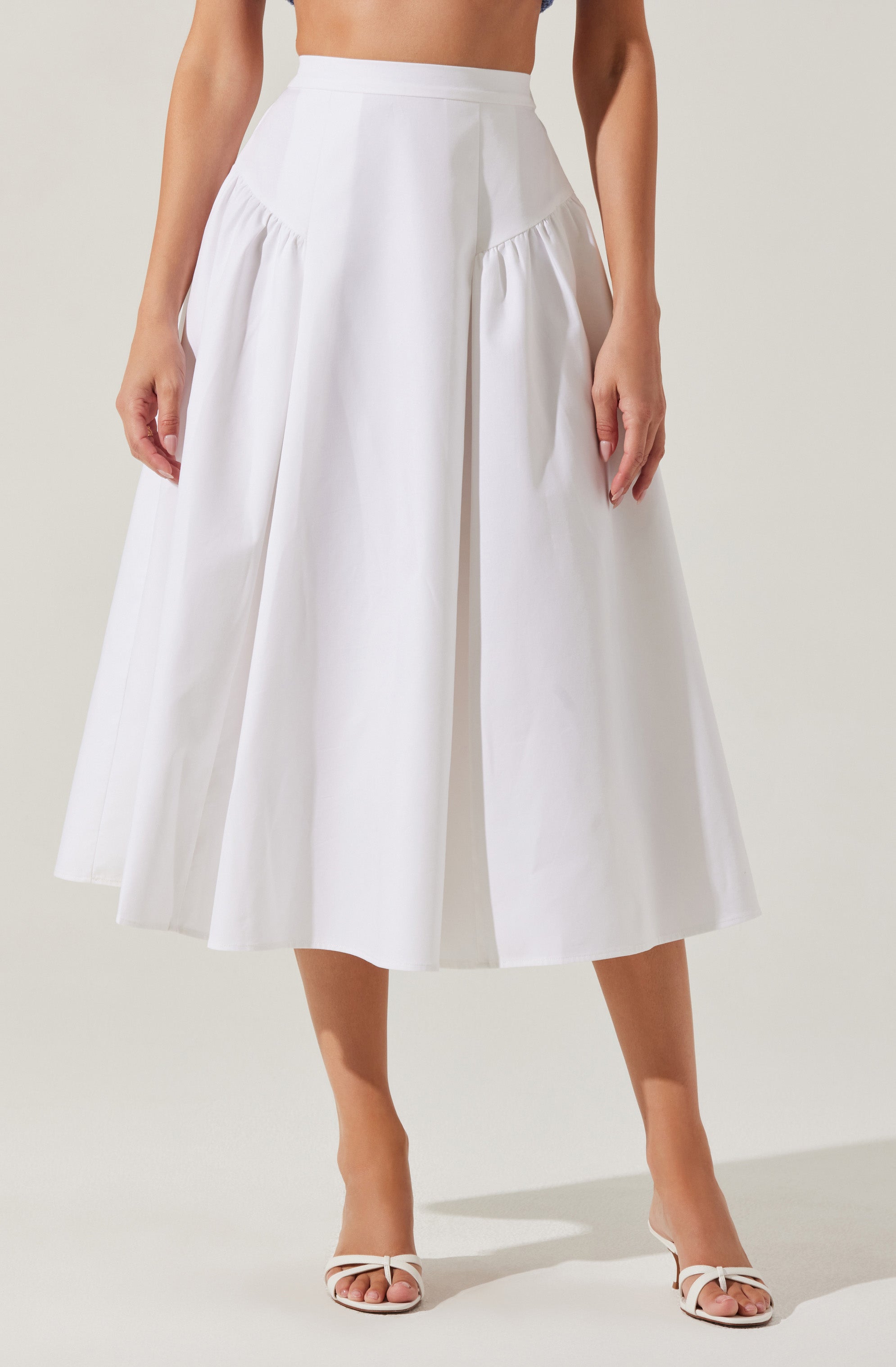 A woman is wearing a high-waisted, white, wide-leg midi skirt with pleats near the waistband. She is also wearing white open-toe heels with ankle straps.