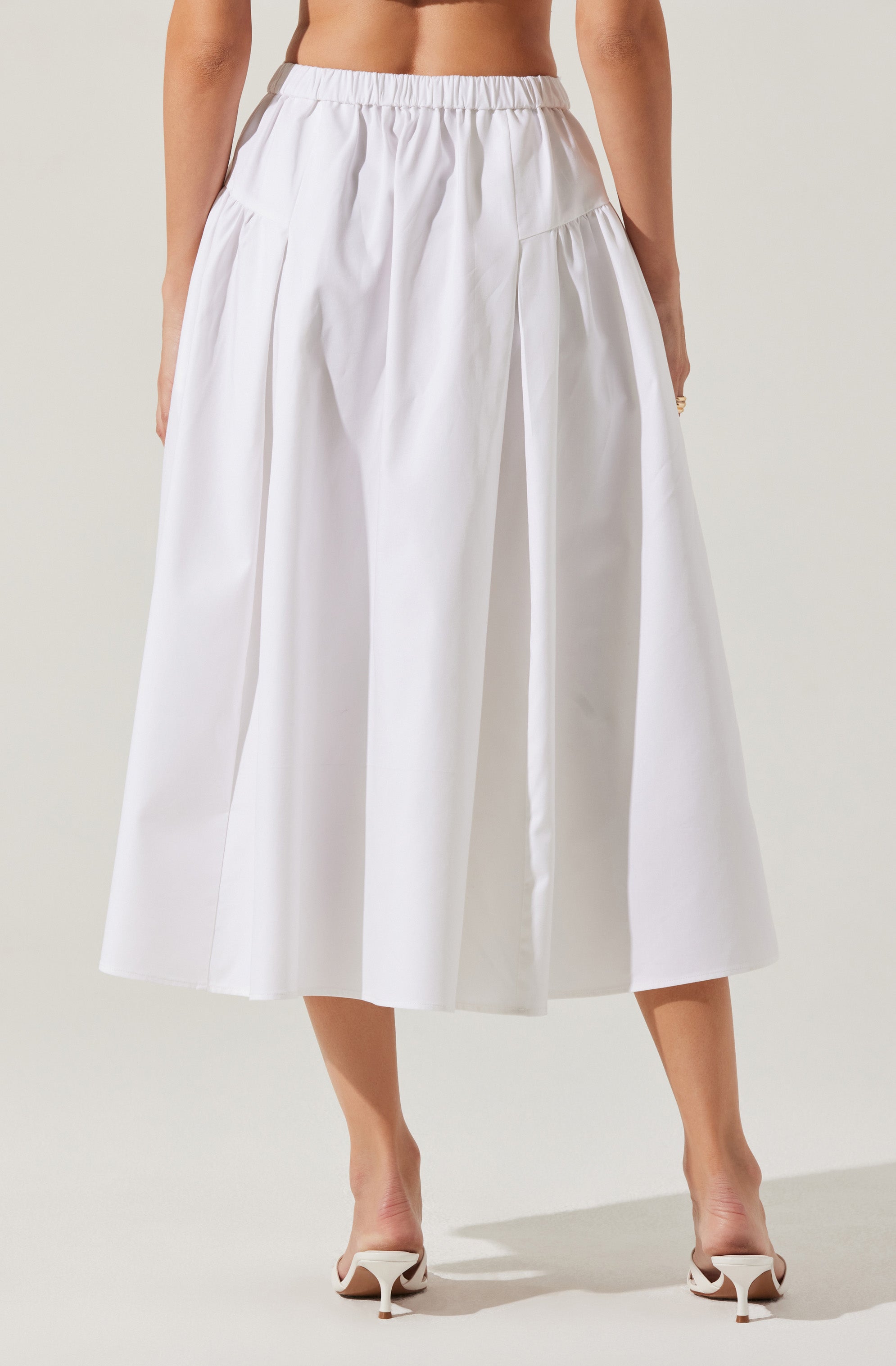 A woman is wearing a white, high-waisted, midi-length skirt with a gathered elastic waistband and pleats that add volume and shape to the skirt. The skirt has a smooth, crisp fabric with a slight sheen. She is also wearing white high-heeled shoes.