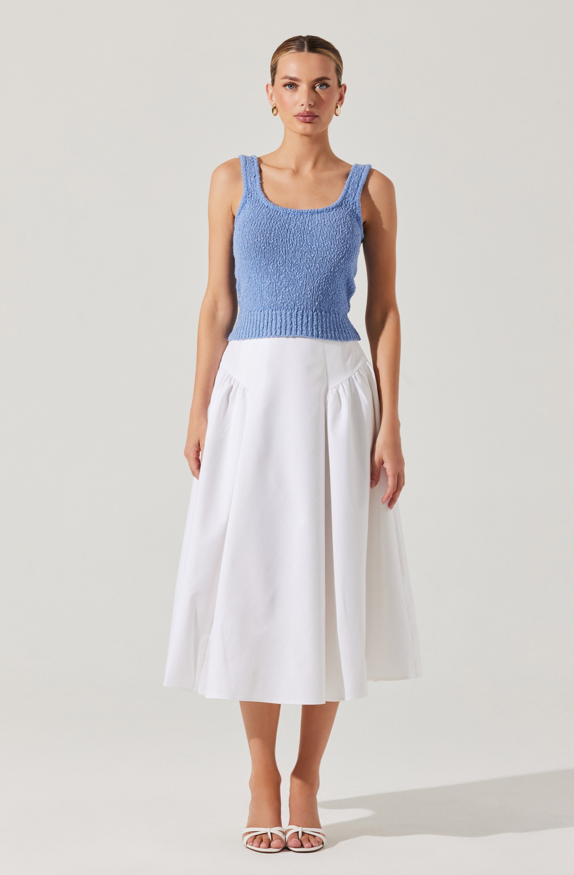 A woman is wearing a sleeveless, light blue knit top with wide straps and a fitted waist. She pairs it with a high-waisted, white, ankle-length skirt that features gathered fabric at the hips and a front slit. She completes her outfit with white strappy high-heeled sandals.