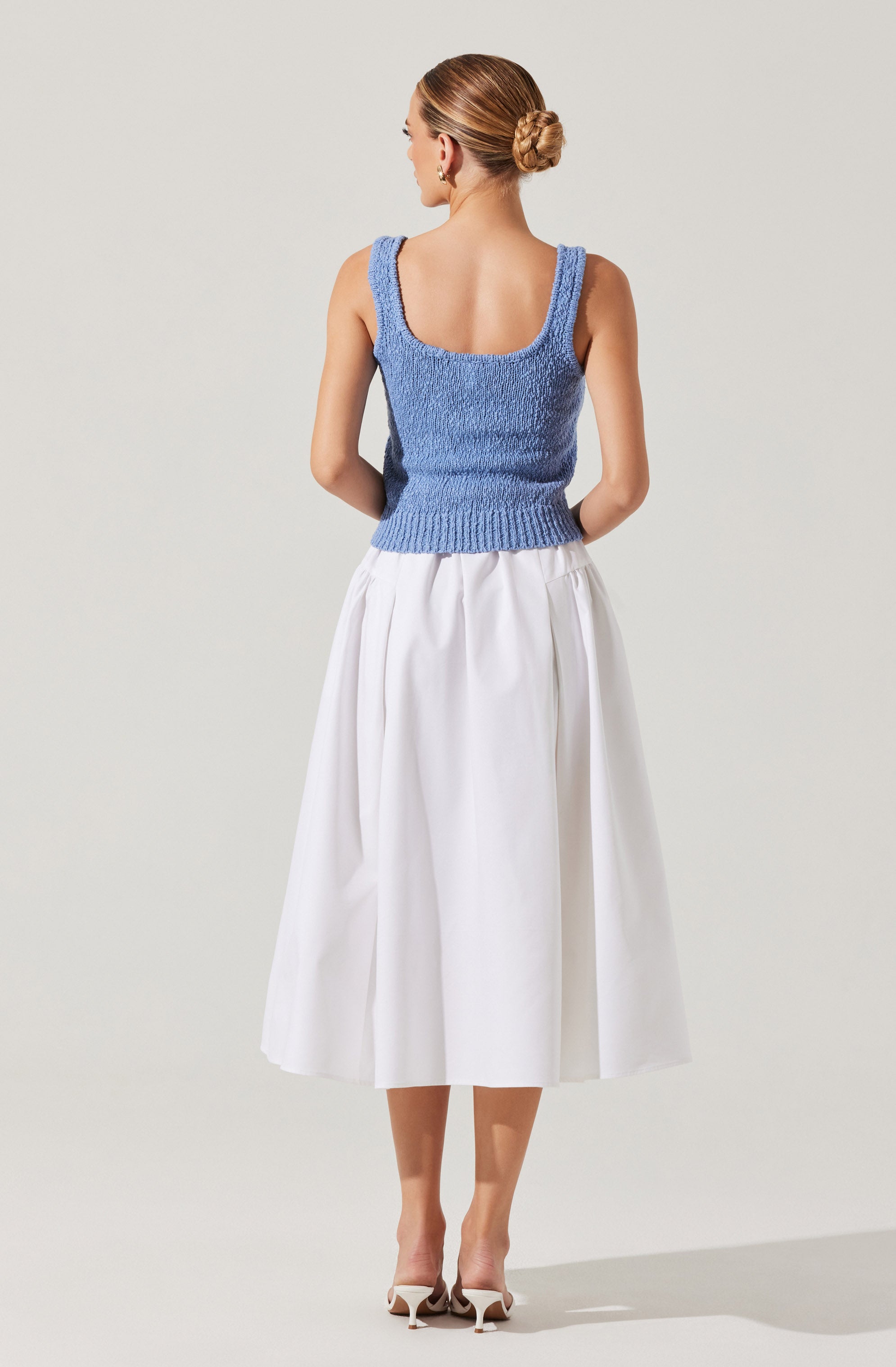 A woman is wearing a sleeveless, textured blue knit top with wide shoulder straps and a square neckline, paired with a high-waisted, full white midi skirt with pleats, and white high-heeled shoes.