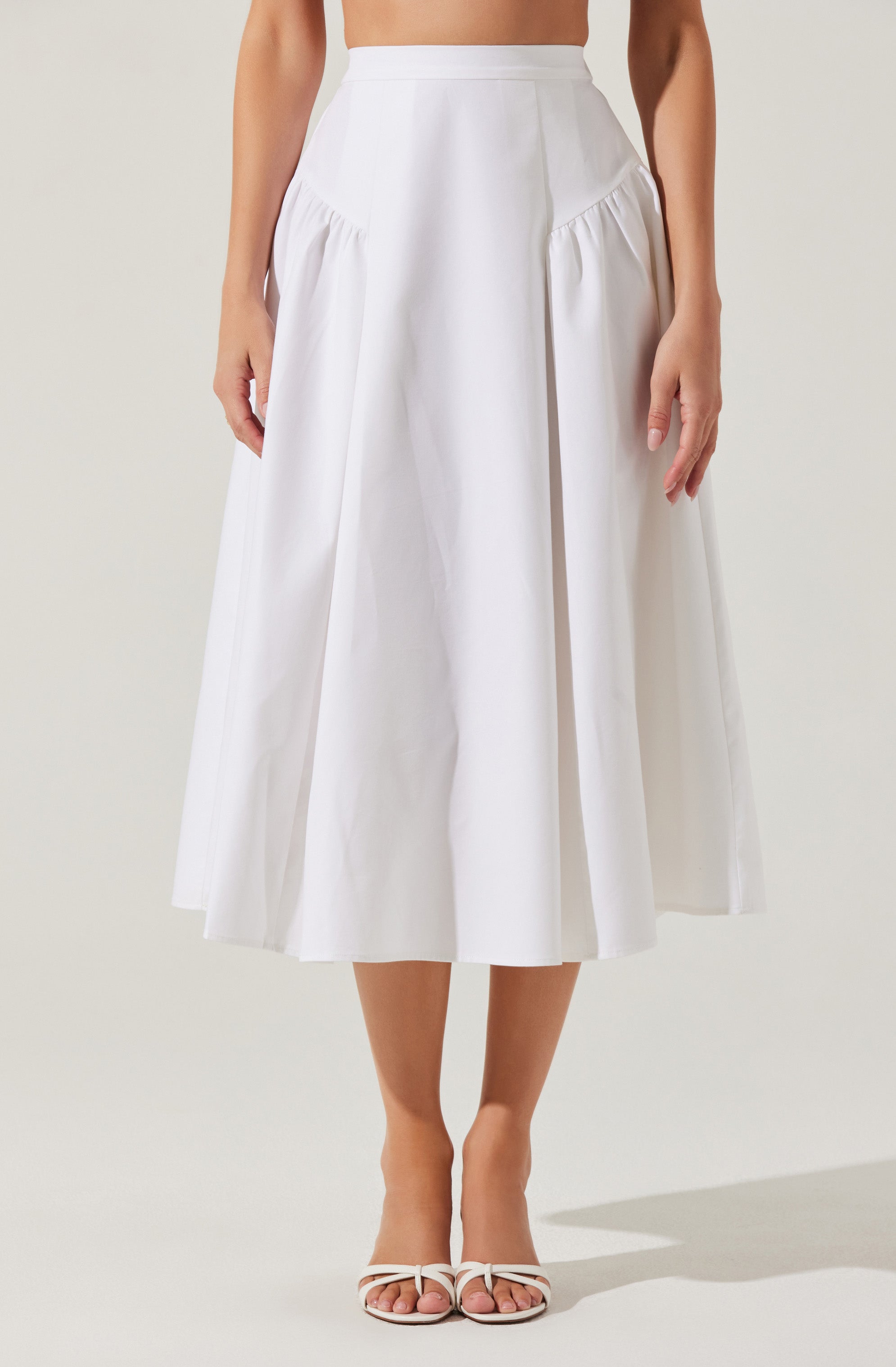 A white, high-waisted, A-line midi skirt with pleats and gathered details on the sides, paired with open-toe white heels.